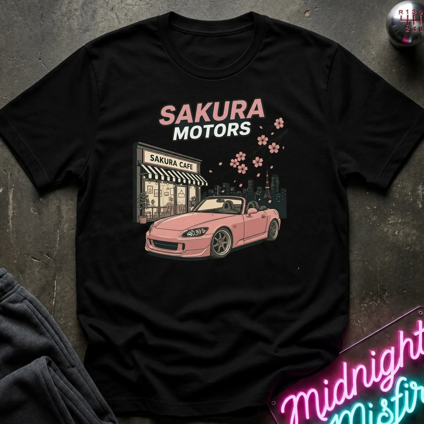A black short-sleeve graphic T-shirt laid flat on a dark textured surface. The shirt features a pink convertible car, a storefront sign reading 'Sakura Cafe', and the words 'SAKURA MOTORS' in pink and white, with small pink cherry blossoms above.