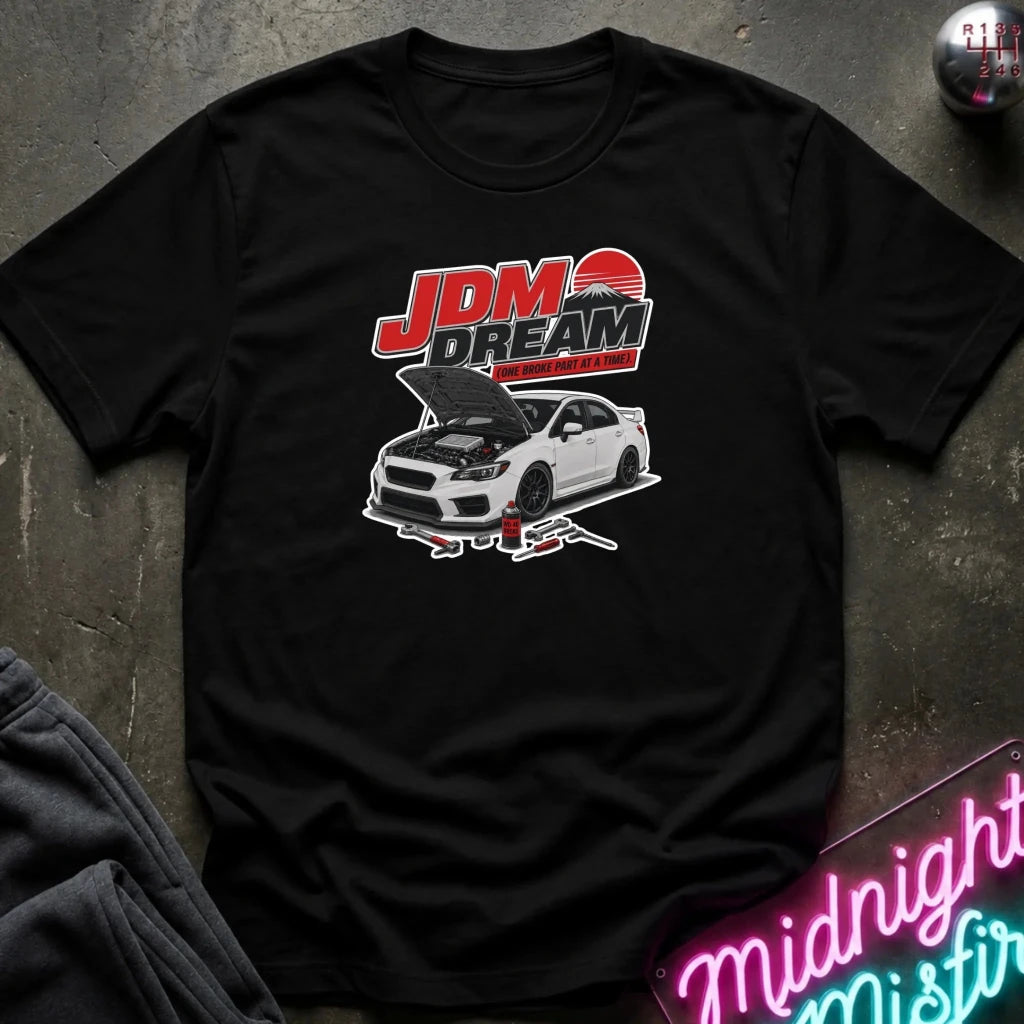 A black short-sleeve graphic t-shirt laid flat on a dark textured surface. The graphic features the text 'JDM DREAM' in red and white above a white sports car with its hood open, supported by jacks.