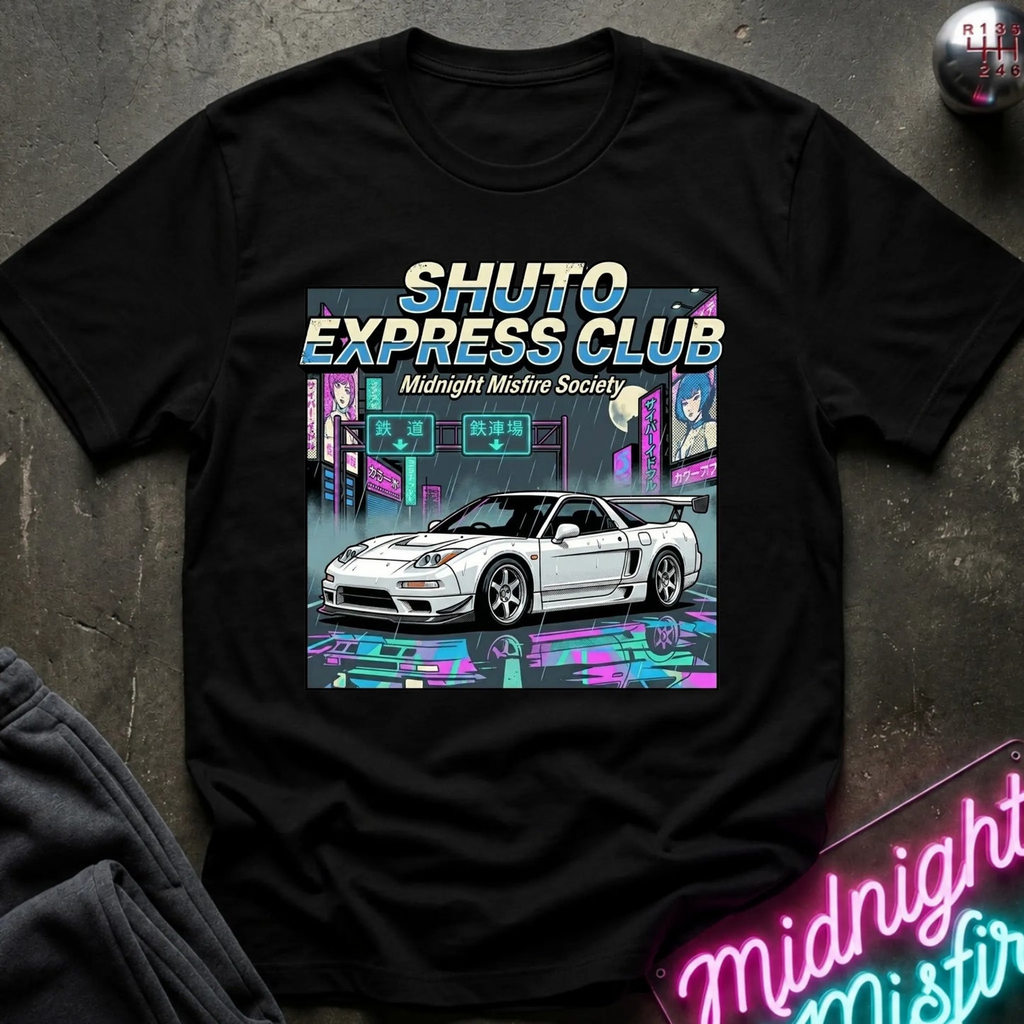 A black short-sleeve graphic T-shirt laid flat on a dark textured surface. The shirt features a large front print with the text 'SHUTO EXPRESS CLUB' and 'Midnight Misfire Society' above an illustration of a white sports car in a neon cyberpunk city scene.