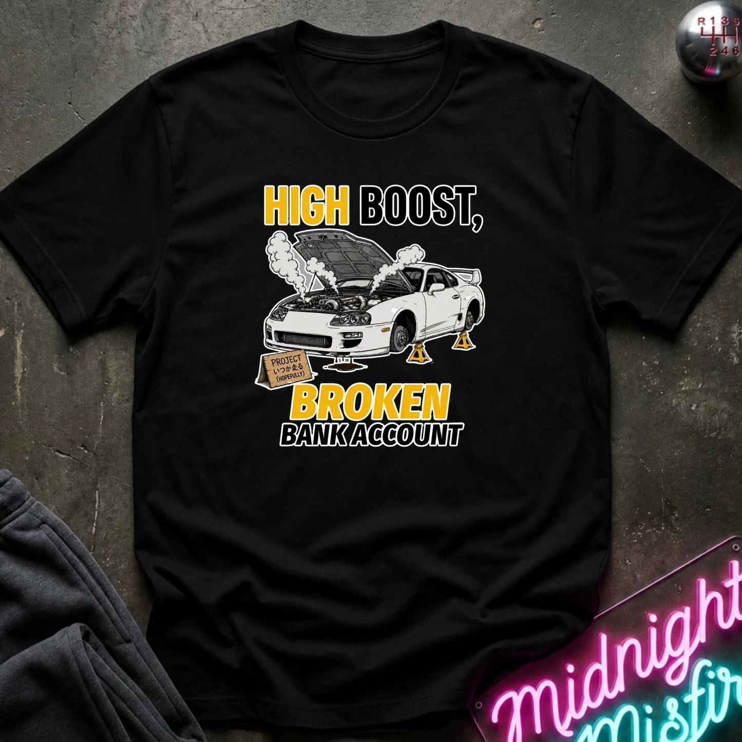 A black short-sleeve graphic T-shirt laid flat on a dark textured surface. The shirt features a white car with its hood open, smoke, and the text 'HIGH BOOST, BROKEN BANK ACCOUNT' in yellow, white, and black.