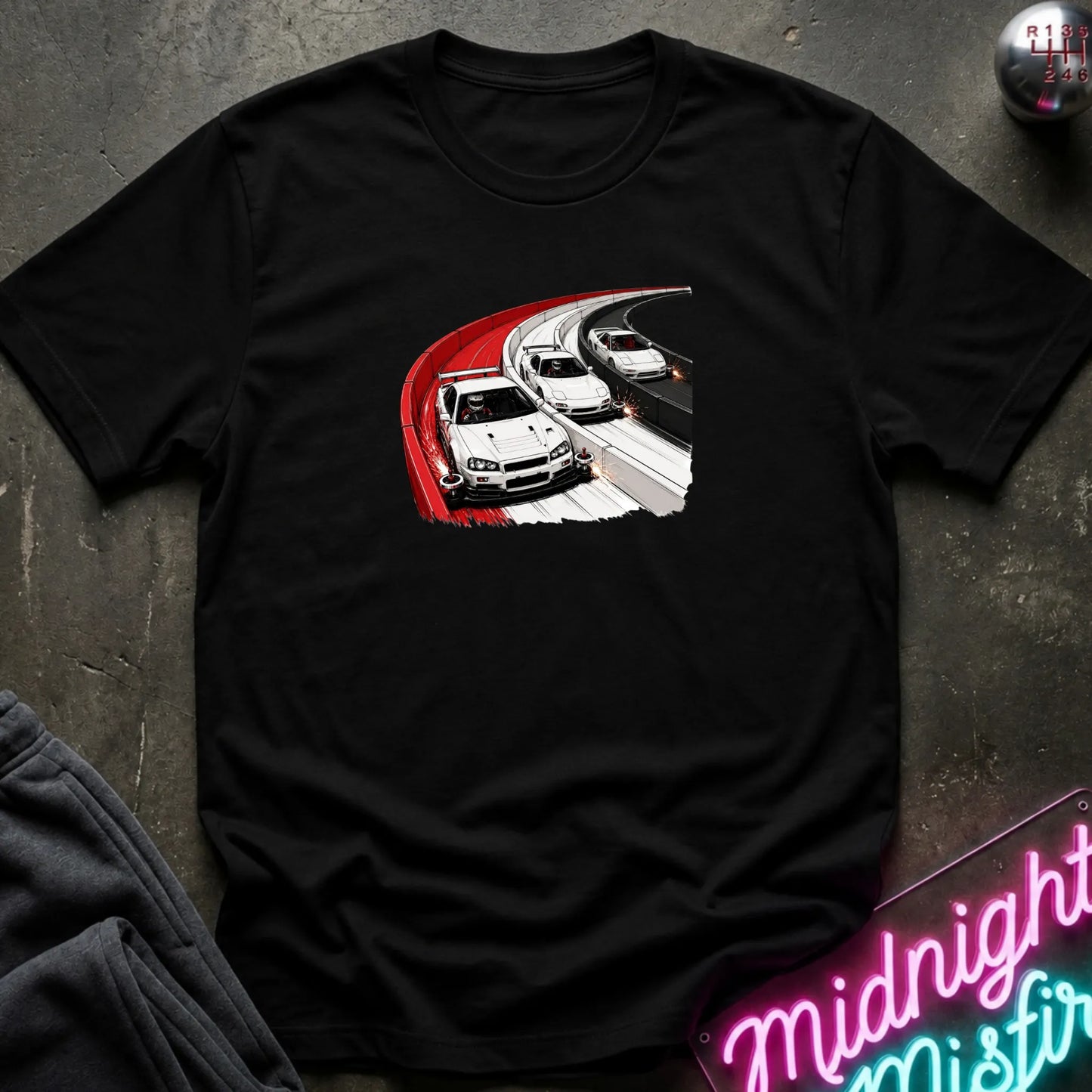 A black short-sleeve t-shirt laid flat on a dark textured surface, featuring a graphic print of three white sports cars racing on a curved road with a red border. 