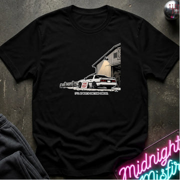 A black short-sleeve crewneck t-shirt laid flat on a dark textured surface, featuring a graphic print of a white sports car parked in front of a barn.