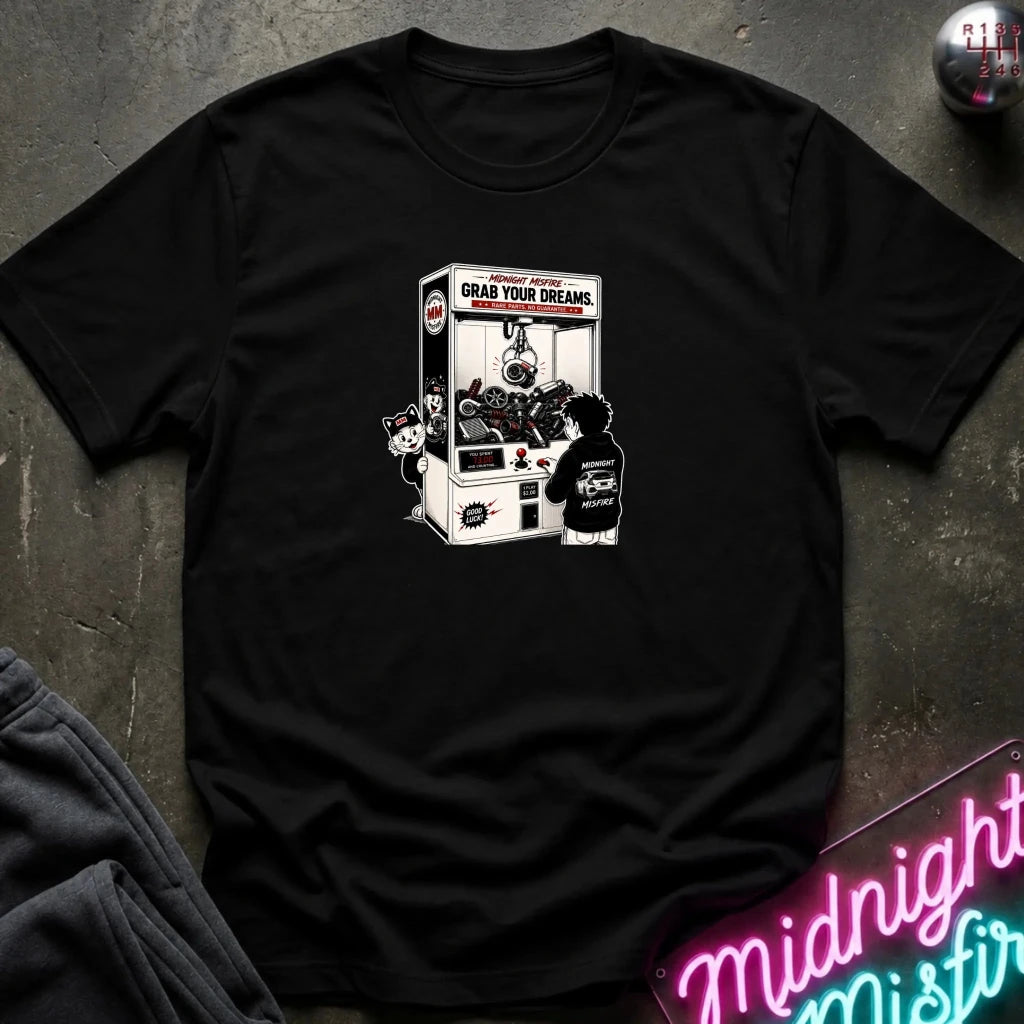 A black short-sleeve t-shirt laid flat on a dark textured surface, featuring a black and white graphic print of a claw machine with the text 'GRAB YOUR DREAMS'. 