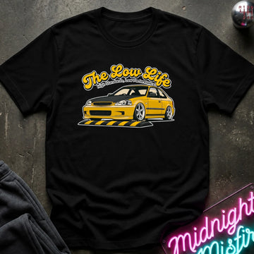 A black short-sleeve graphic t-shirt laid flat on a dark textured surface. The shirt features a yellow and white graphic of a yellow car with the text 'The Low Life' in yellow script.
