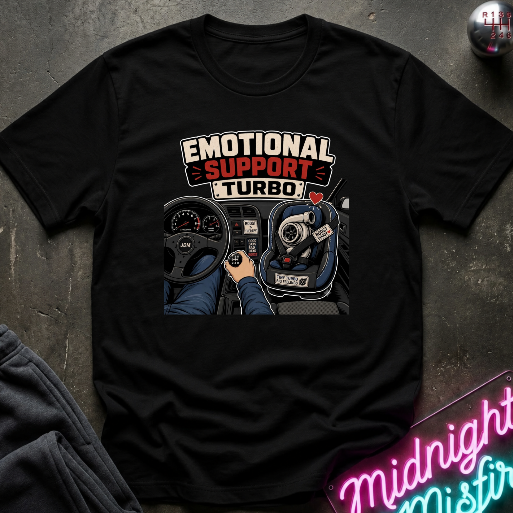 A black short-sleeve graphic t-shirt laid flat on a dark textured surface. The shirt features a graphic print with the words 'EMOTIONAL SUPPORT TURBO' above an illustration of a hand on a steering wheel inside a car.