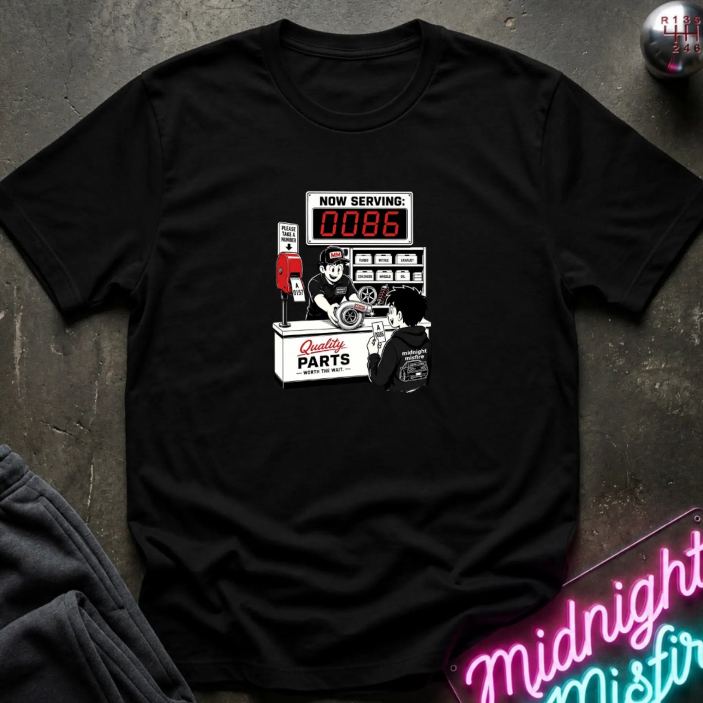 A black  short-sleeve t-shirt with a retro-style graphic of a garage attendant at a 'Quality Parts' counter, displayed flat on a dark textured surface with a neon sign and sweatpants partially visible.