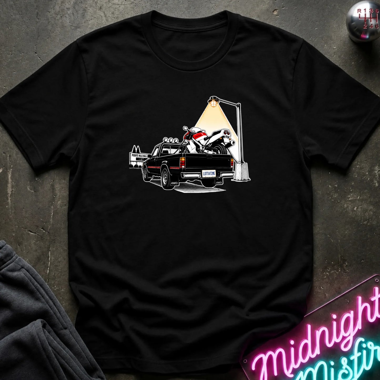 A black short-sleeve crewneck t-shirt featuring a graphic print of a black pickup truck carrying a white and red motorcycle under a street lamp, laid flat on a dark textured surface.