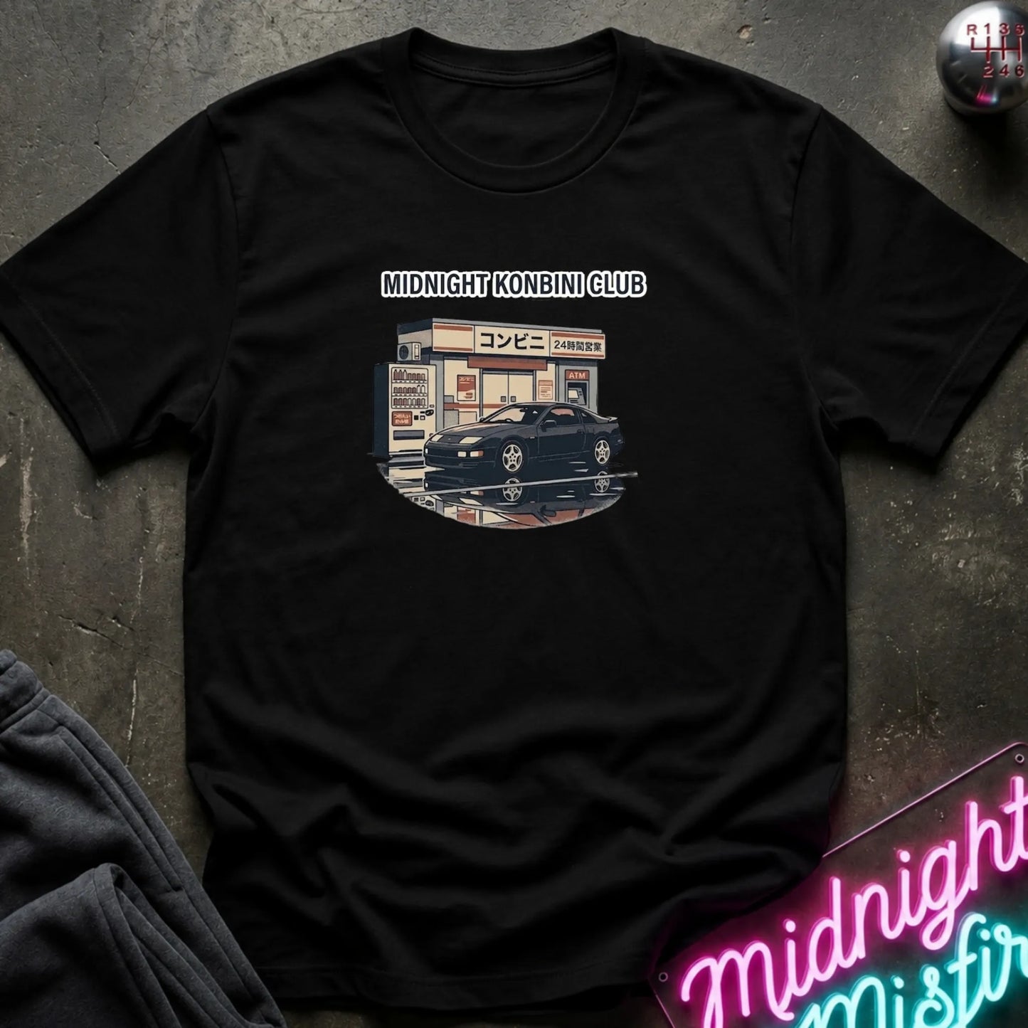 A black short-sleeve t-shirt laid flat on a dark textured surface, featuring a graphic print of a black sports car parked in front of a convenience store with the text 'MIDNIGHT KONBINI CLUB' above it. A neon sign and folded dark fabric are partially visible around the shirt.
