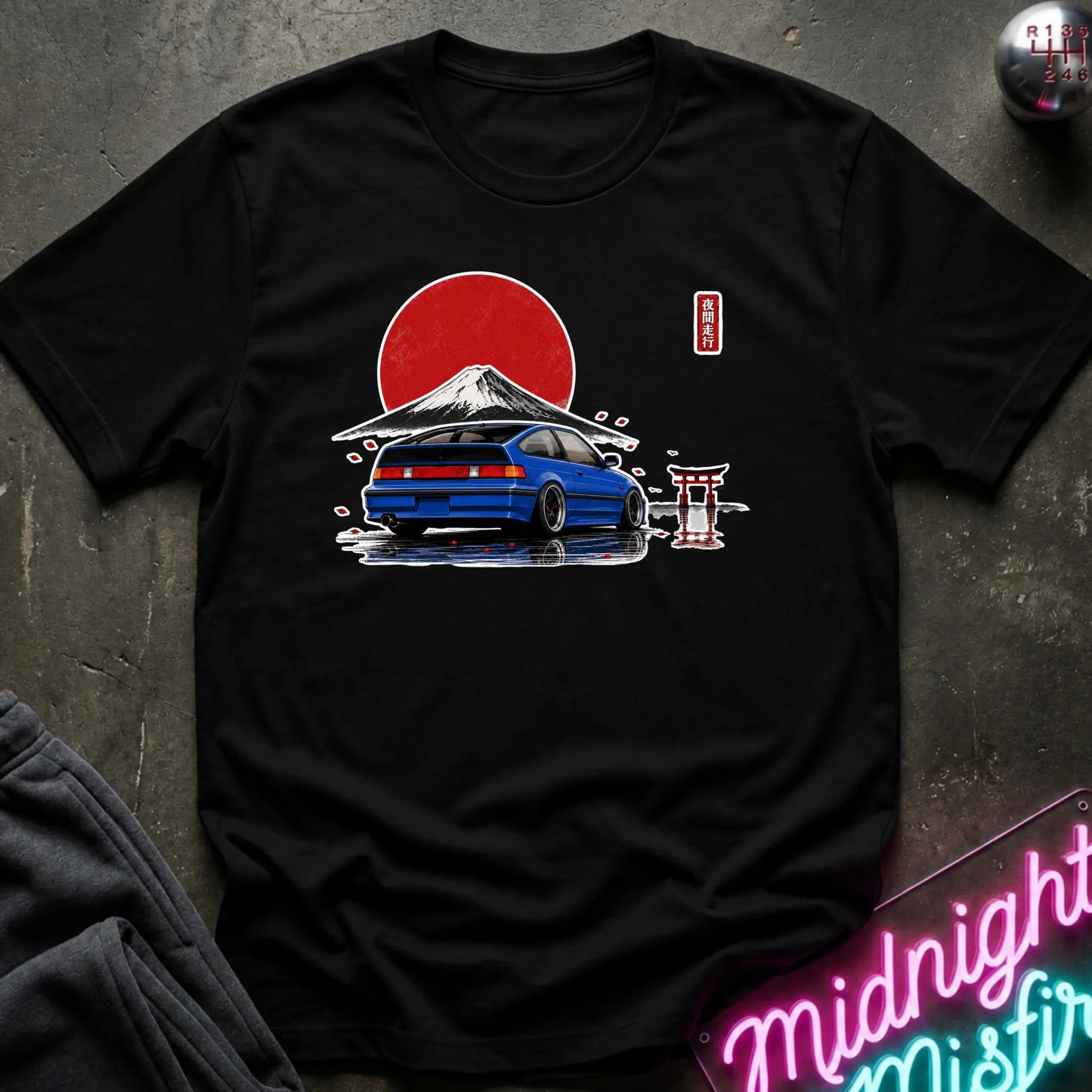 A black short-sleeve graphic t-shirt laid flat on a dark textured surface. The graphic features a blue sports car in front of a red sun and a snow-capped mountain, with a small red vertical logo on the upper right chest.