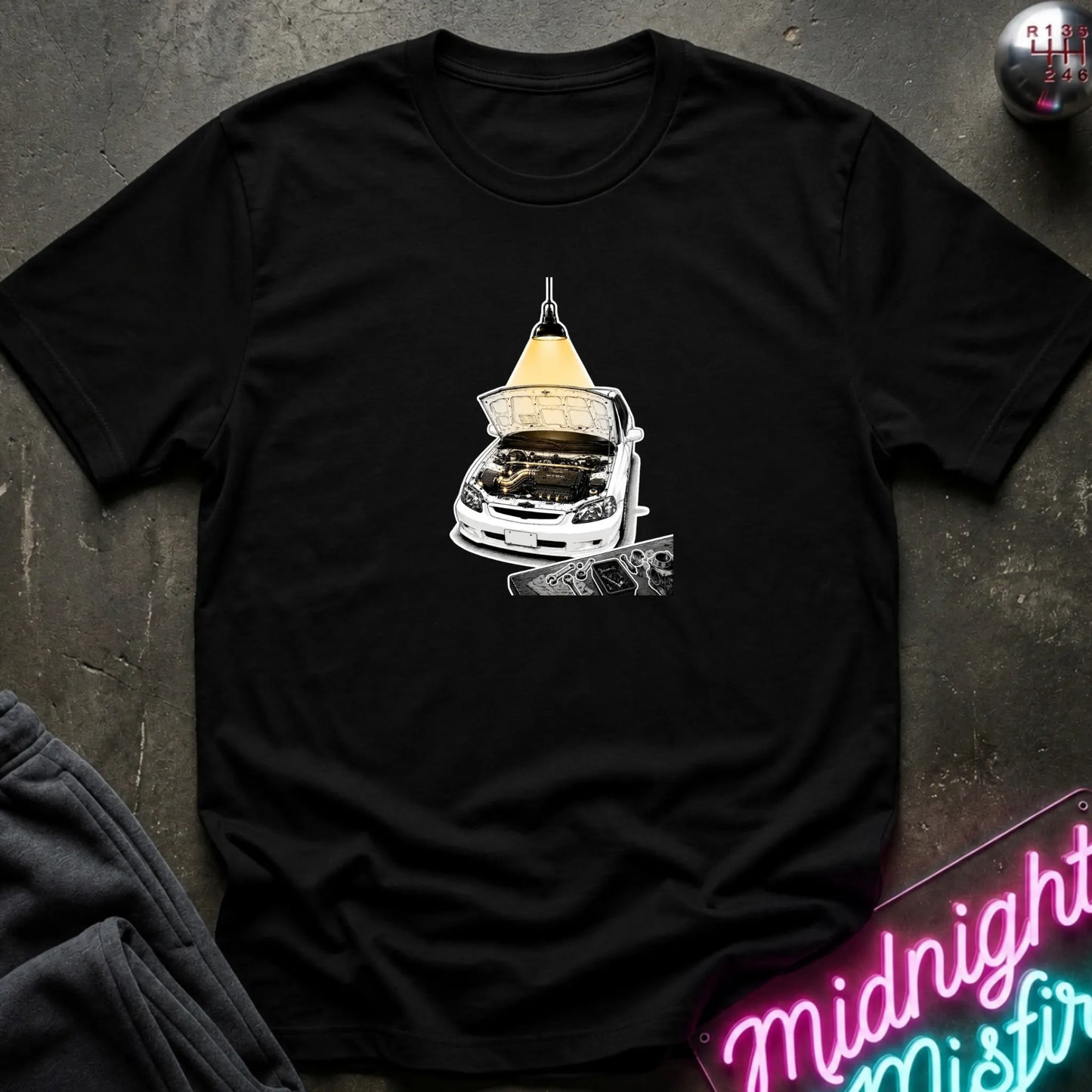 A black short-sleeve t-shirt laid flat on a dark textured surface, featuring a graphic print of a white car with its hood open under a glowing yellow lamp. 