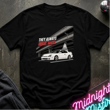A black short-sleeve graphic t-shirt laid flat on a dark textured surface. The graphic features a white sports car under a bridge with the text 'THEY ALWAYS COME BACK' in white and red.