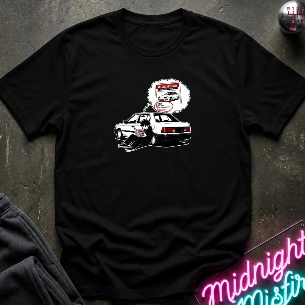 A black short-sleeve t-shirt laid flat on a dark textured surface, featuring a graphic of a black-and-whitecar with a thought bubble showing a car part 