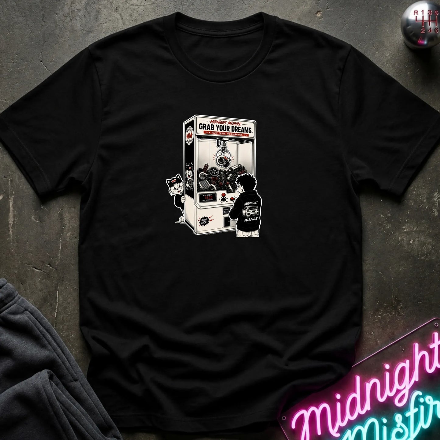 A black short-sleeve crewneck t-shirt laid flat on a dark textured surface, featuring a central graphic of a person in a black hoodie playing a claw machine. 