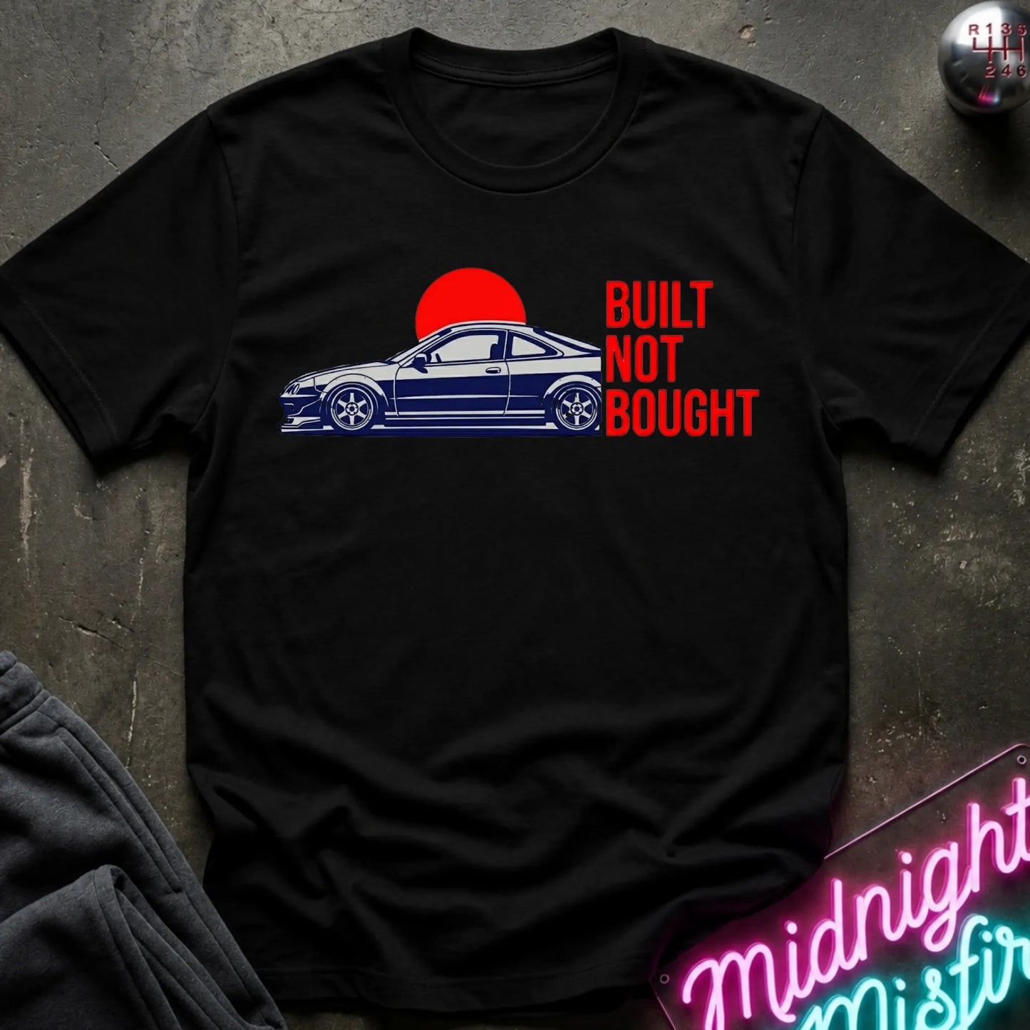 A black short-sleeve graphic T-shirt laid flat on a dark textured surface. The shirt features a blue side-profile car graphic under a red sun and the phrase 'BUILT NOT BOUGHT' in red text.