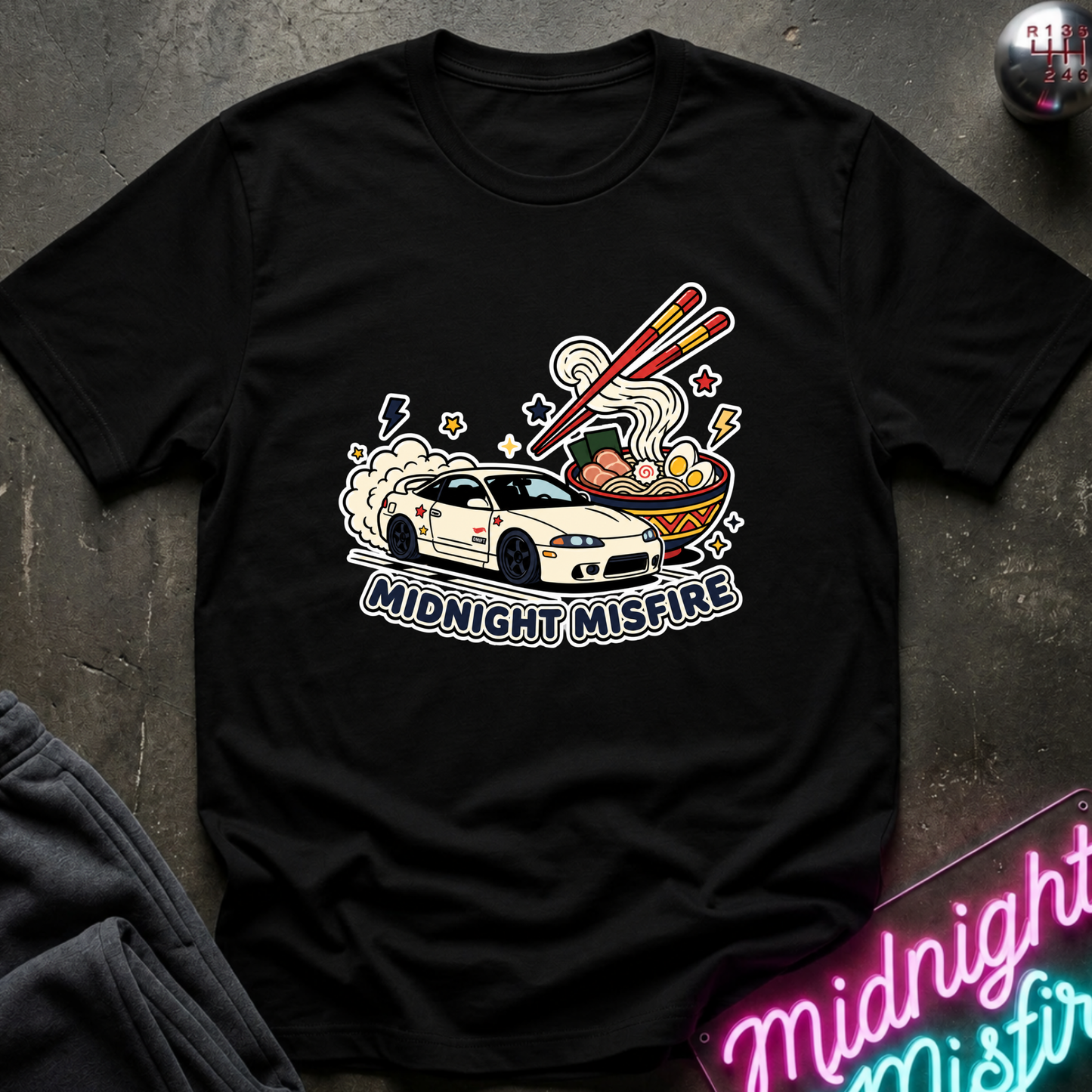 A black short-sleeve graphic t-shirt laid flat on a dark textured surface. The graphic features a white race car, chopsticks, ramen noodles, and the text 'MIDNIGHT MISFIRE'.
