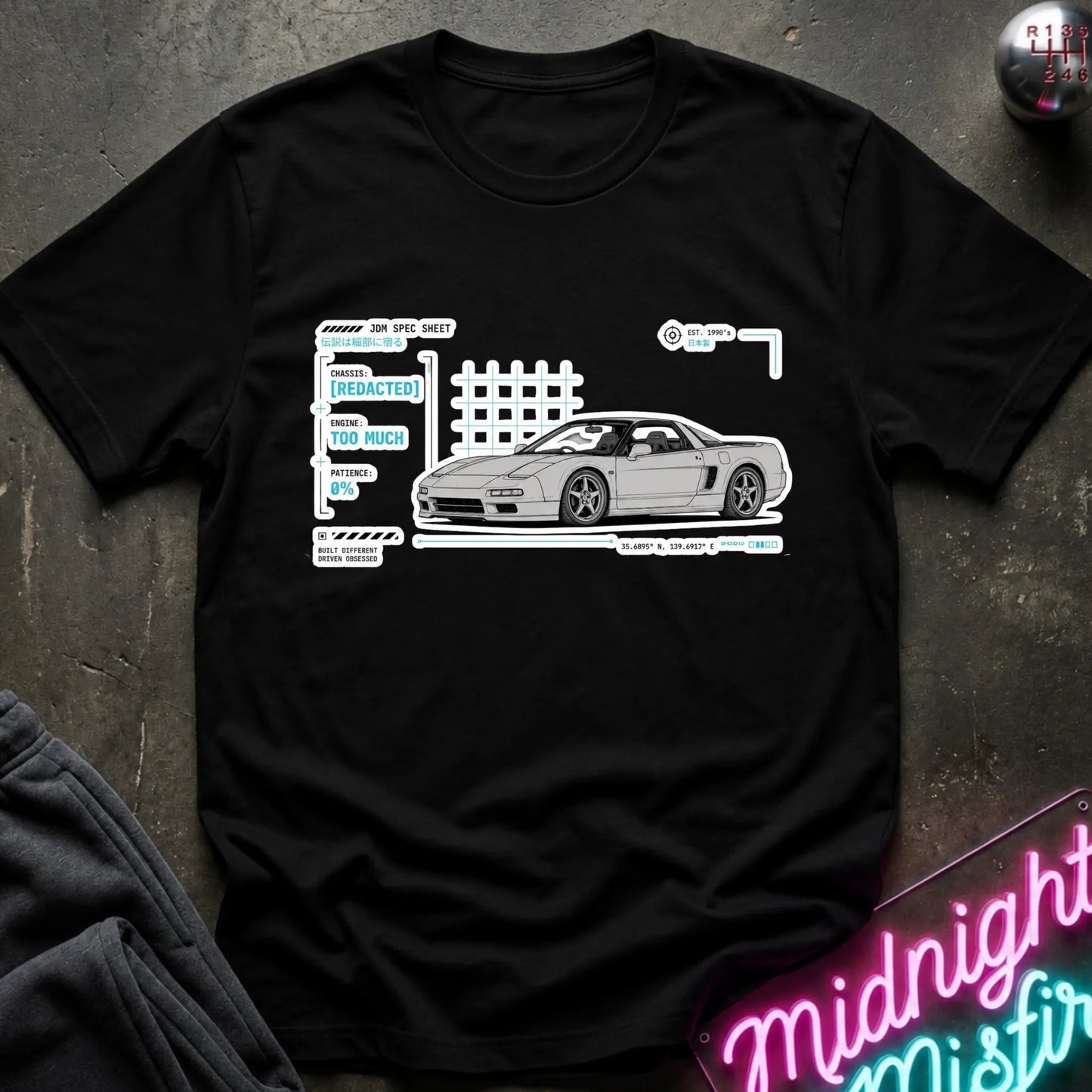 A black short-sleeve t-shirt laid flat on a dark textured surface, featuring a white and light blue graphic print of a sports car with technical-style labels. A neon sign and folded dark fabric are partially visible around the edges.
