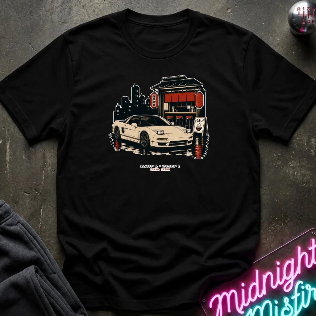 A black short-sleeve graphic T-shirt laid flat on a dark textured surface. The shirt features a central graphic of a white sports car parked in front of a red and beige building with a city skyline in the background.