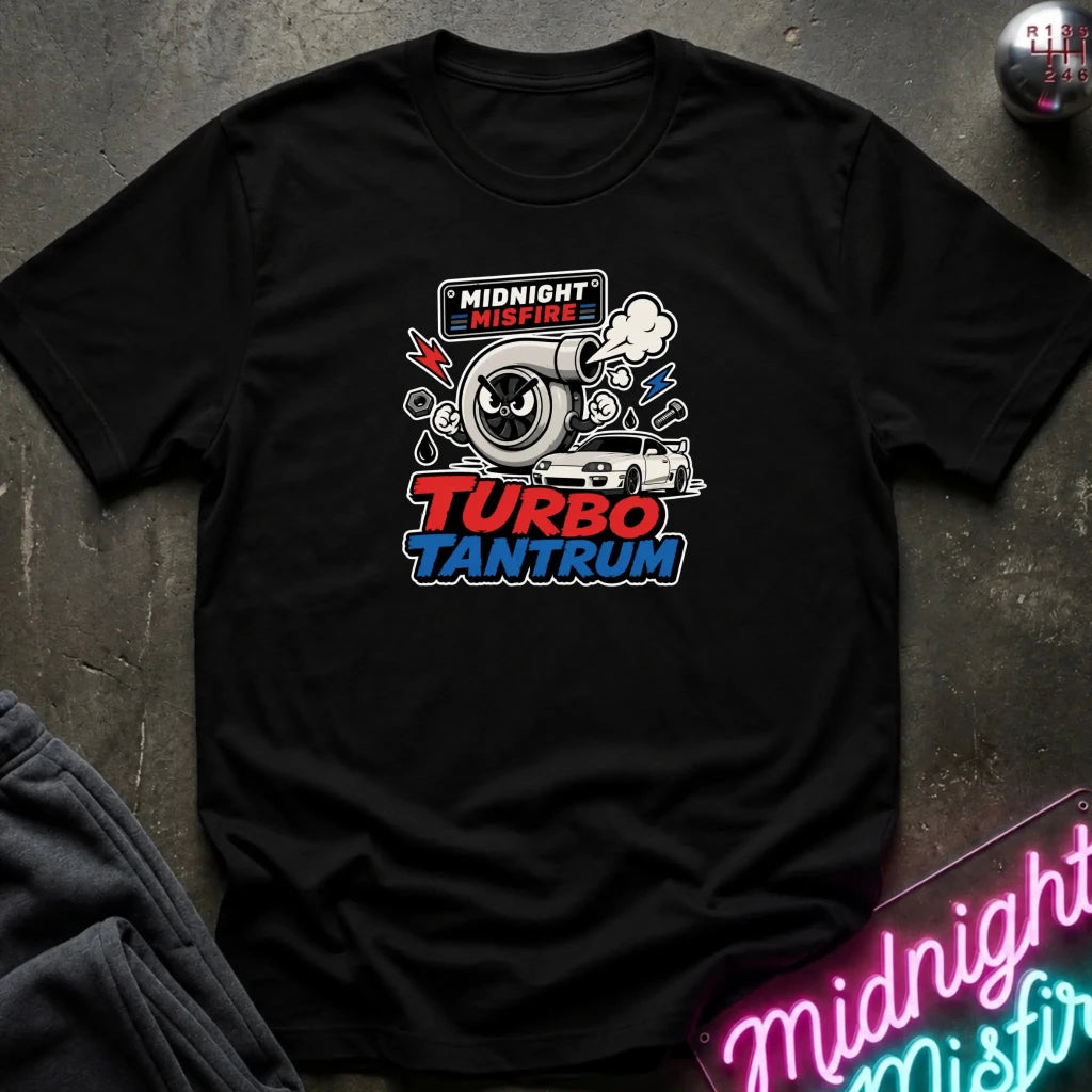 A black short-sleeve graphic t-shirt laid flat on a dark textured surface. The shirt features a central graphic of a turbocharger, a white car, and the text 'MIDNIGHT MISFIRE' and 'TURBO TANTRUM' in red, white, and blue.