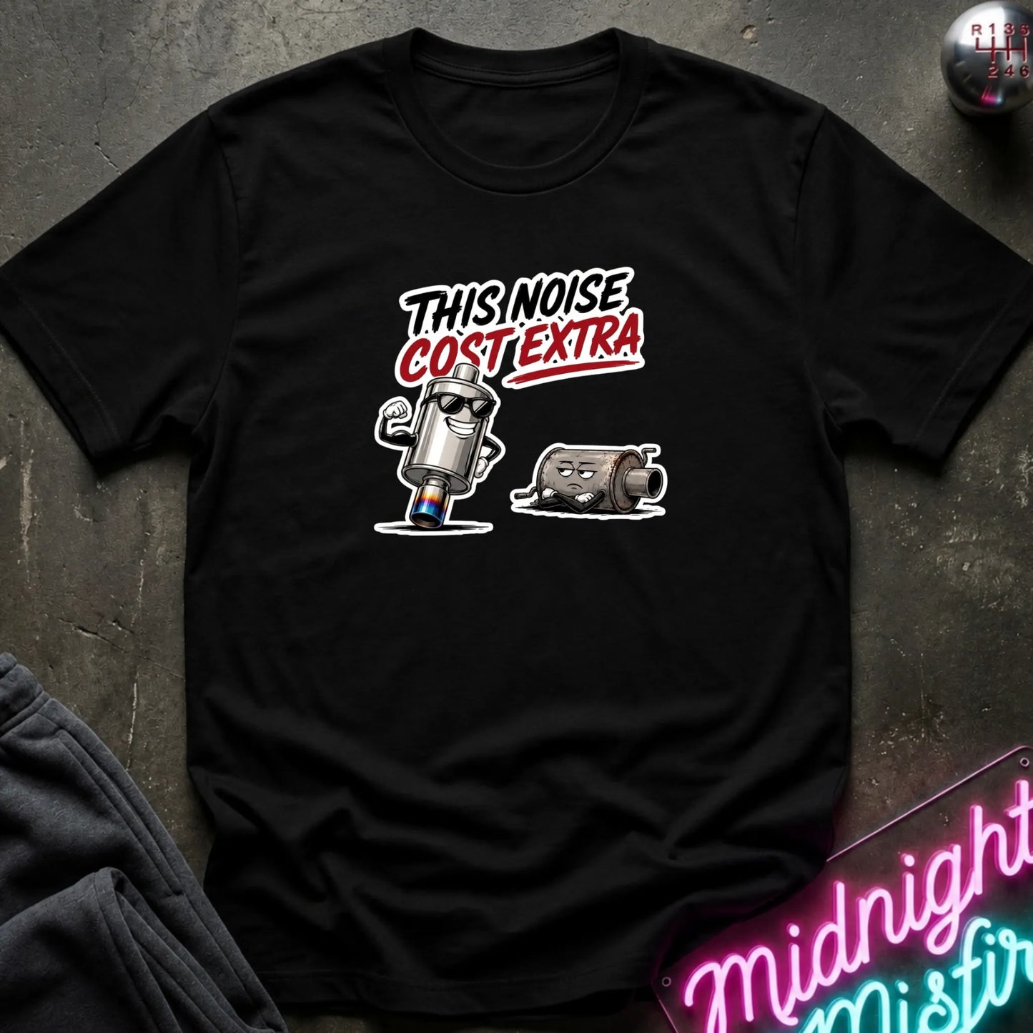 A black short-sleeve graphic t-shirt laid flat on a dark textured surface. The graphic features the text 'THIS NOISE COST EXTRA'.