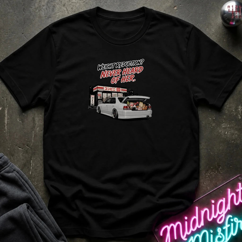 A black short-sleeve graphic t-shirt laid flat on a dark textured surface. The graphic features a white station wagon with its trunk open, parked in front of a gas station, with the text 