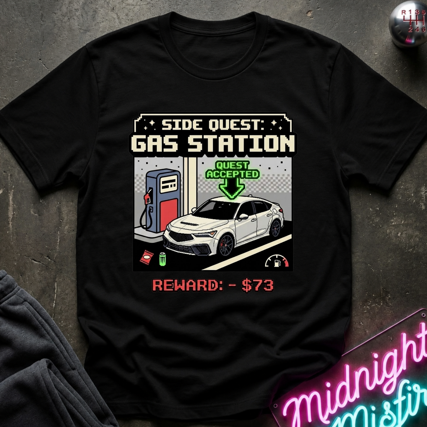 A black short-sleeve graphic t-shirt laid flat on a dark textured surface. The shirt features a retro video game style print with the text 'SIDE QUEST: GAS STATION', an illustration of a white car at a gas pump, and 'GUEST ACCEPTED' with a green arrow.