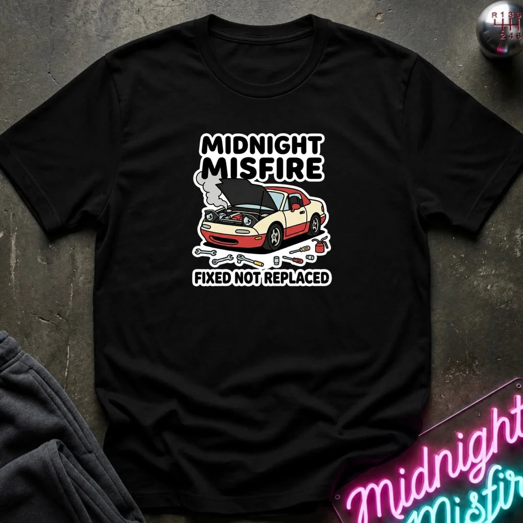 A black short-sleeve graphic T-shirt laid flat on a dark textured surface. The shirt features a white and red graphic of a car with smoke and the text 'MIDNIGHT MISFIRE' above and 'FIXED NOT REPLACED' below.