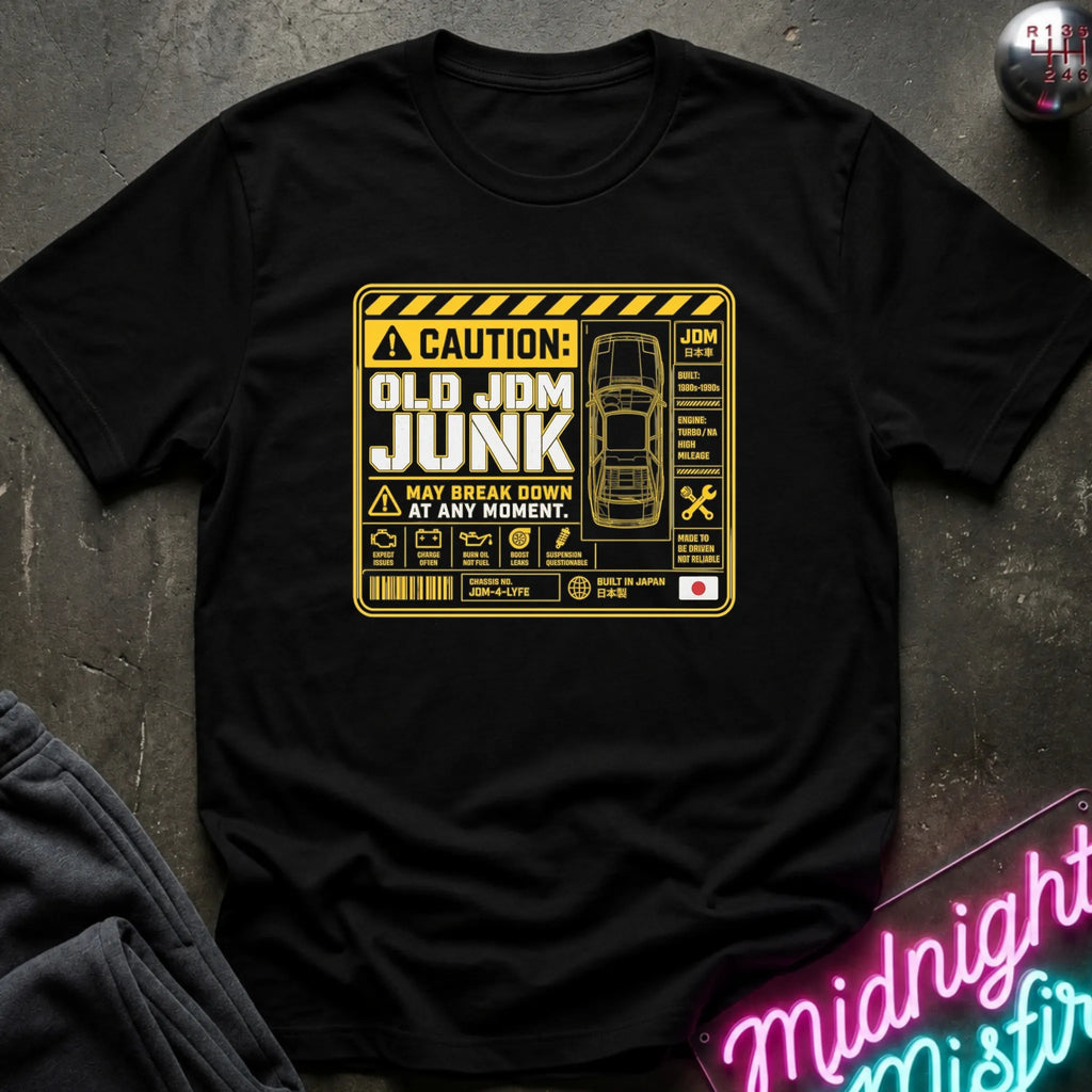 A black short-sleeve graphic T-shirt laid flat on a dark textured surface. The shirt features a large yellow caution-style print reading 'OLD JDM JUNK' with a car outline and technical-style icons.