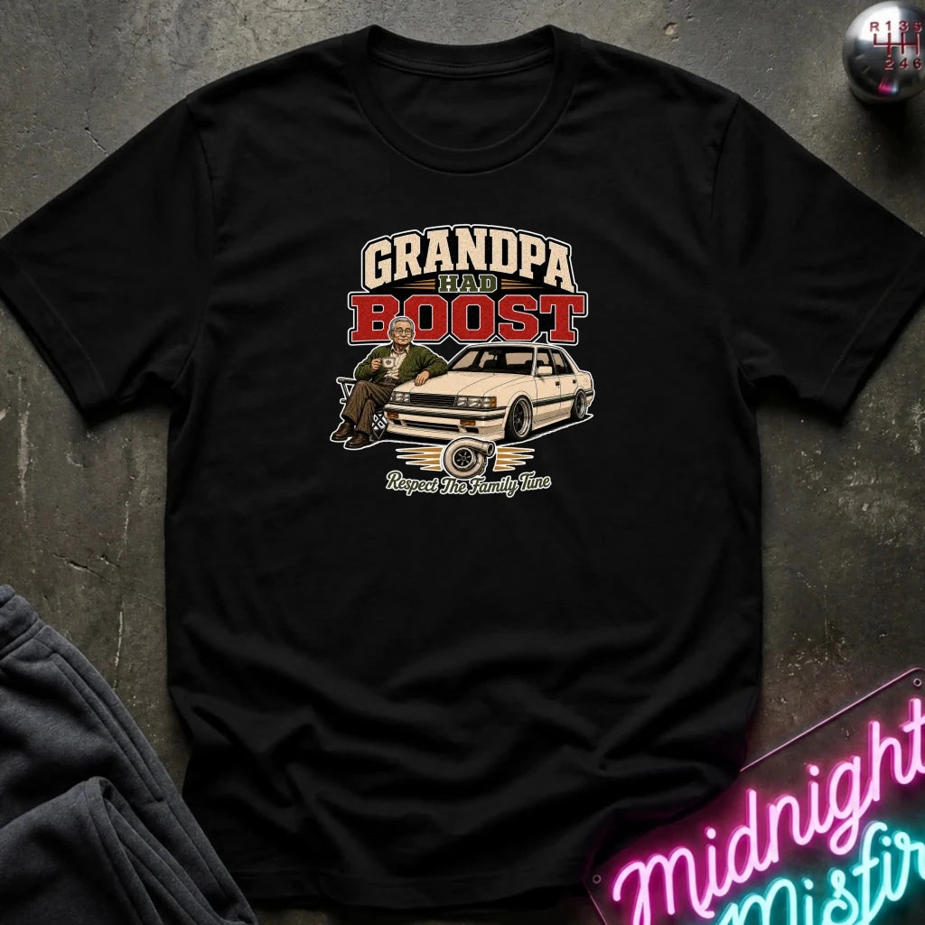black short-sleeve graphic t-shirt laid flat on a dark textured surface. The graphic features the text 'GRANDPA RAW BOOST' in beige and red, above an illustration of a man sitting in a car with the phrase 'Respect The Family Tune' below.
