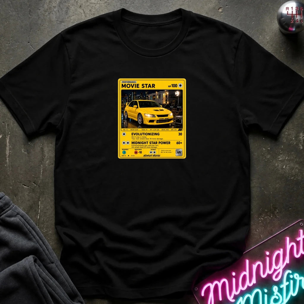 A black short-sleeve t-shirt laid flat on a dark textured surface, featuring a yellow and black retro-style graphic on the chest with a yellow car photo and text including 'MOVIE STAR'. 