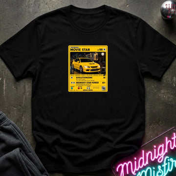 A black short-sleeve t-shirt laid flat on a dark textured surface, featuring a yellow and black retro-style graphic on the chest with a yellow car photo and text including 'MOVIE STAR'. 