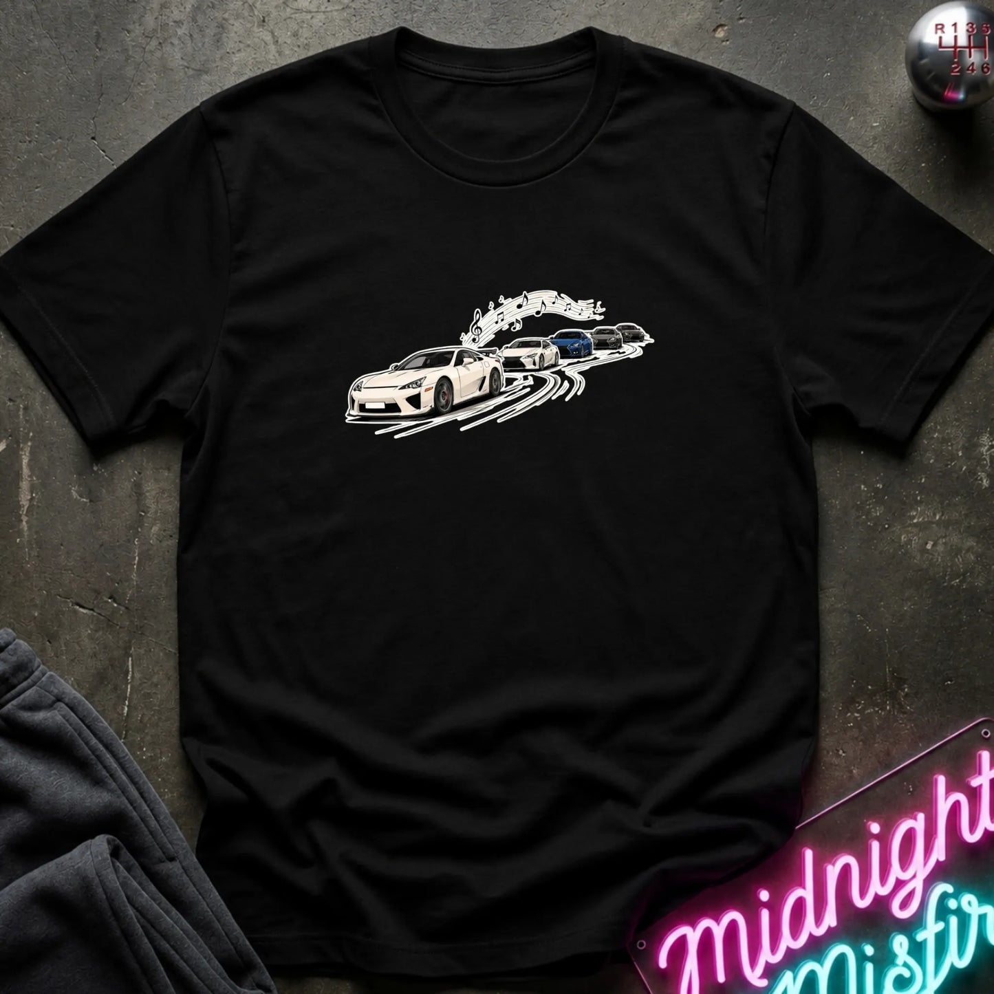A black short-sleeve crewneck t-shirt featuring a graphic print of a white sports car leading a line of other cars, laid flat on a dark textured surface with a neon sign and dark pants partially visible.
