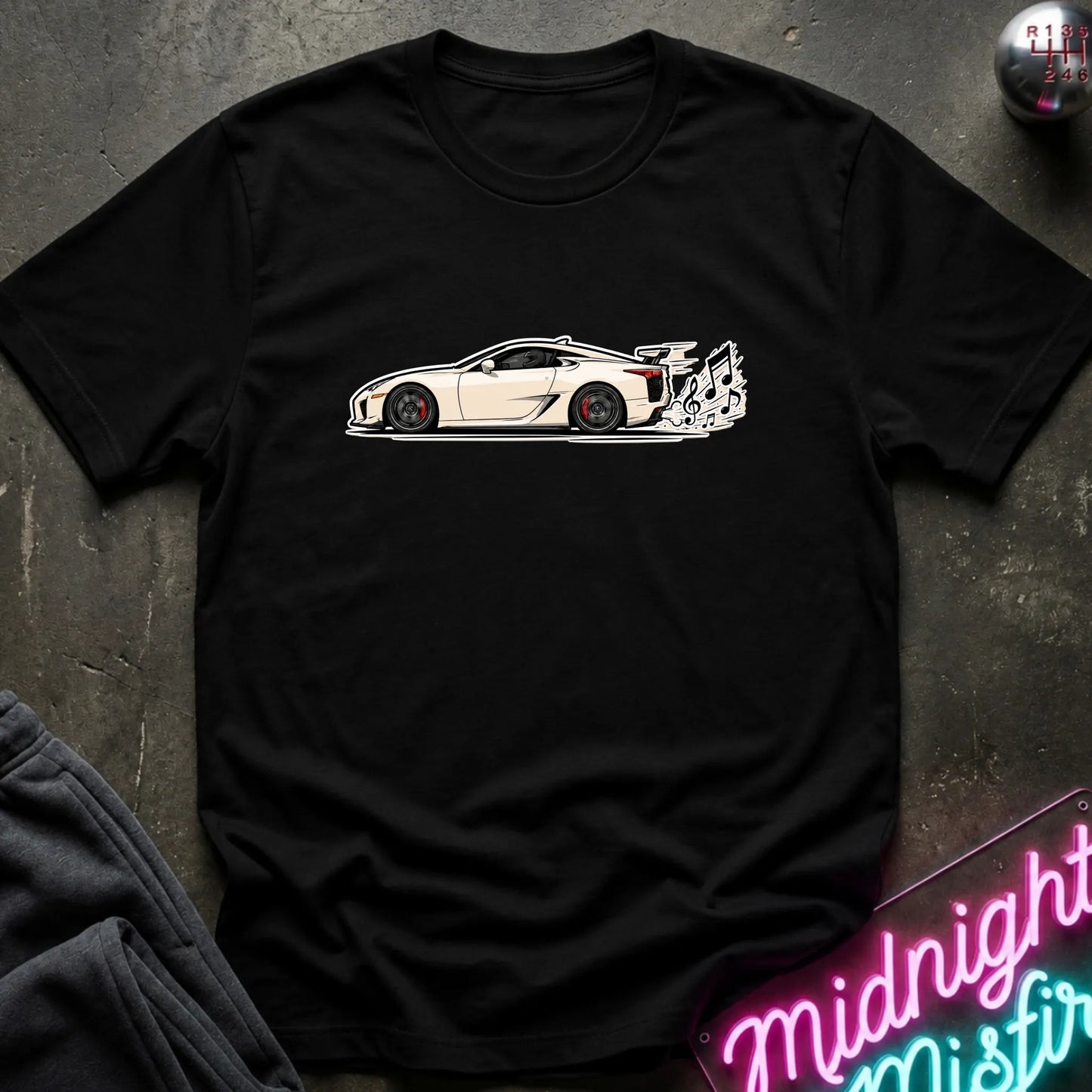 A black short-sleeve t-shirt laid flat on a dark textured surface, featuring a graphic print of a white sports car with a music note coming from exhaust.