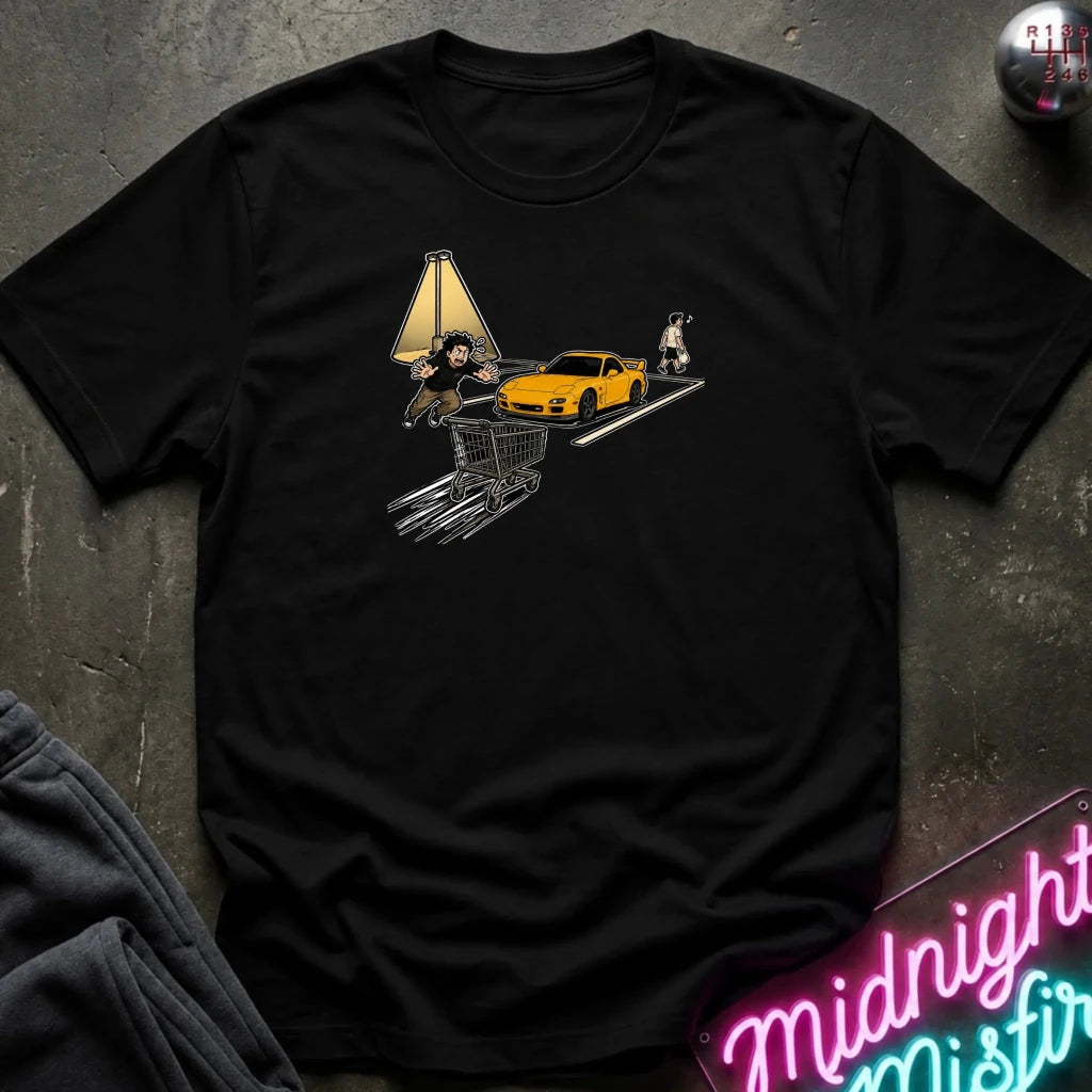 black crewneck t-shirt laid flat on a dark textured surface. The shirt features a graphic print of a yellow sports car, a shopping cart, a lamp, and a person, styled with a dark sweatshirt and a neon sign in the corners.