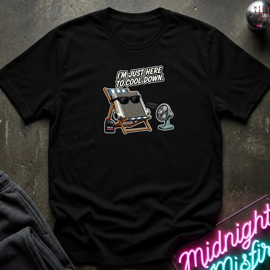 A black short-sleeve graphic t-shirt laid flat on a dark textured surface. The graphic features a vintage slide-rule calculating machine on a beach chair with sunglasses, a small fan, and the text 'I'M JUST HERE TO COOL DOWN.
