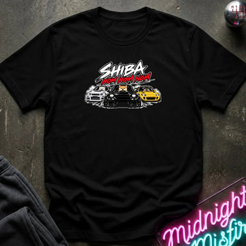 A black short-sleeve graphic t-shirt laid flat on a dark textured surface. The graphic features the text 'SHIBA NOM NOM NOM' in white and red, above an illustration of three cars (white, black, and yellow).