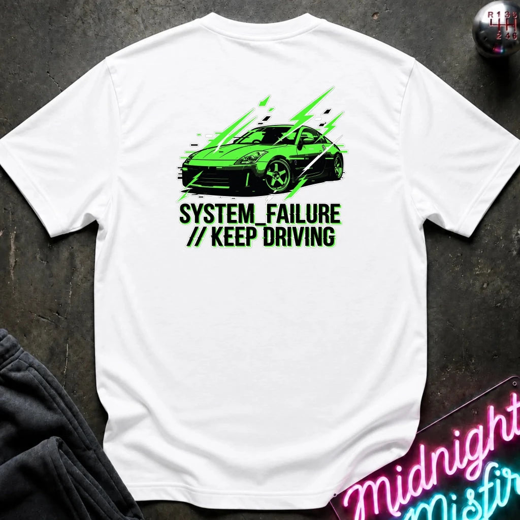 white short-sleeve t-shirt laid flat on a dark textured surface, featuring a graphic of a green sports car with text 'SYSTEM FAILURE // KEEP DRIVING' in green. A neon sign and dark clothing are partially visible as props.