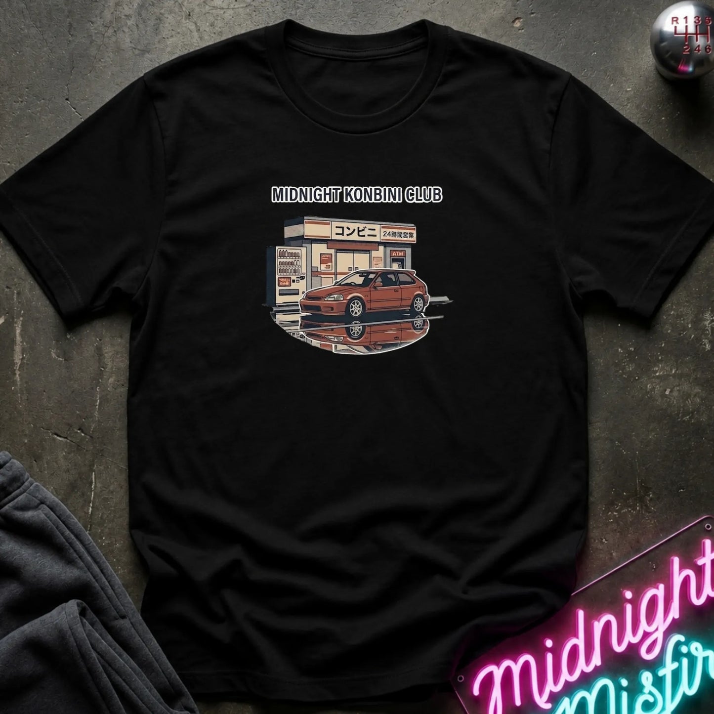 A black short-sleeve t-shirt laid flat on a dark textured surface, featuring a graphic print of a red sports car parked in front of a convenience store with the text 'MIDNIGHT KONBINI CLUB' above it. A neon sign and dark clothing are partially visible as props around the shirt.