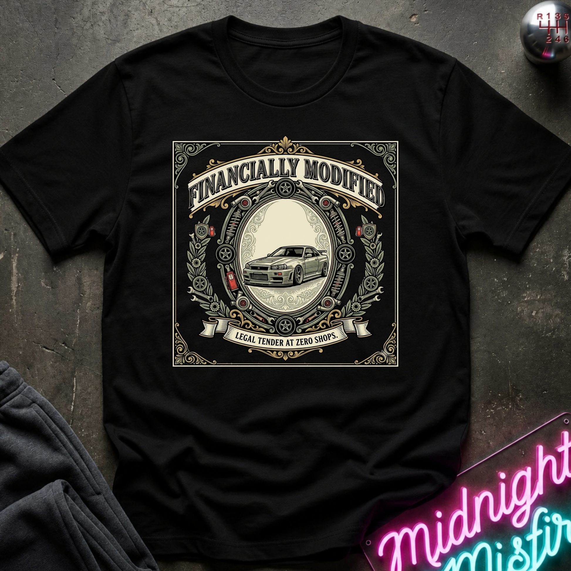 A black short-sleeve graphic t-shirt laid flat on a dark textured surface. The shirt features a large vintage-style circular graphic with the text 'FINANCALLY MODIFIED' and 'LOCAL TENDER AT ZERO SHOPS' surrounding an illustration of a classic car.