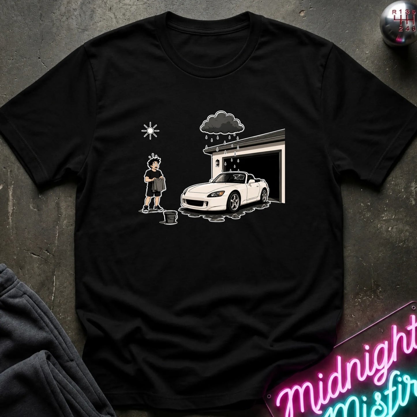 A black short-sleeve crewneck t-shirt laid flat on a dark textured surface. The shirt features a white graphic print of a person washing a white sports car in front of a garage under a dark raincloud.