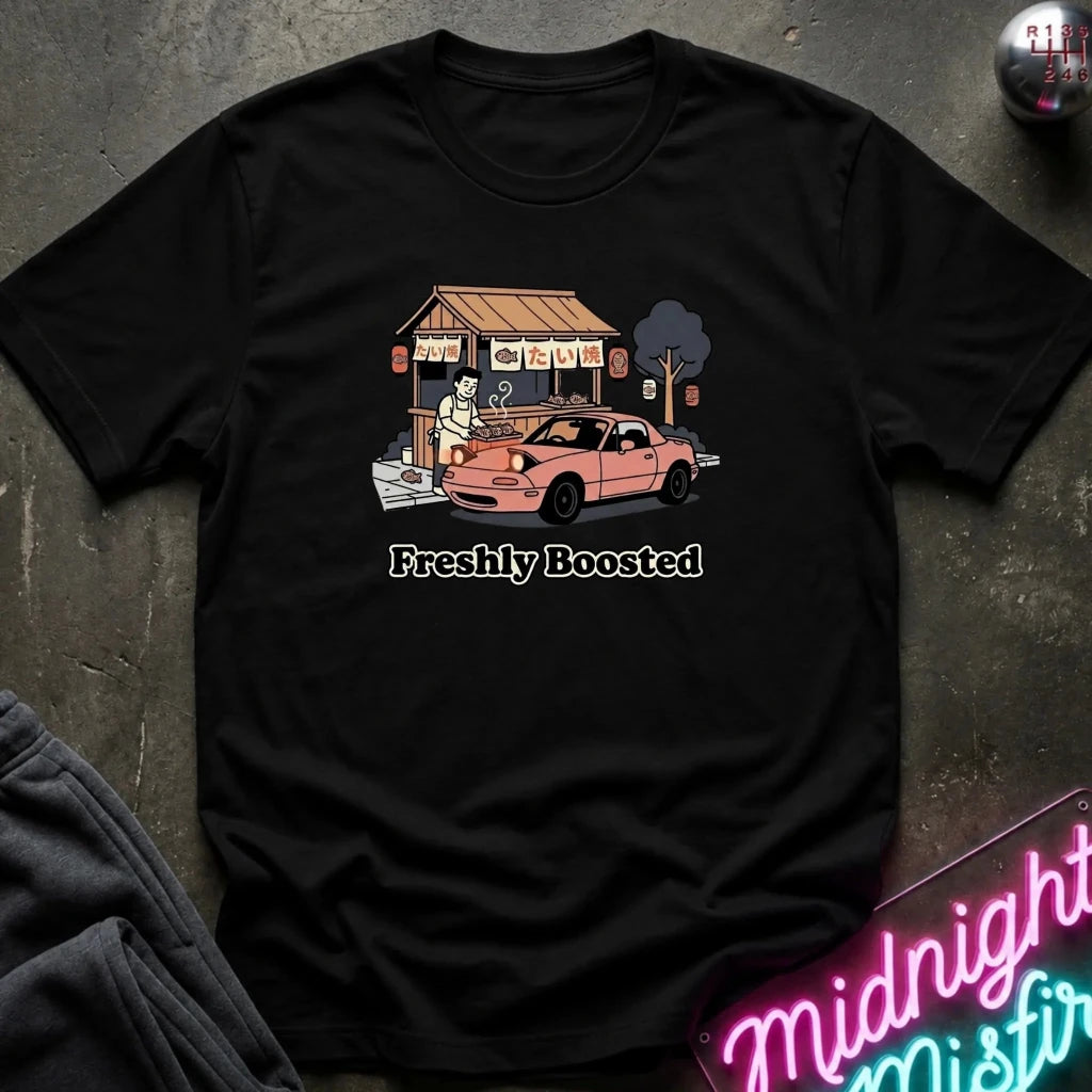 A black short-sleeve graphic t-shirt laid flat on a dark textured surface. The graphic shows a pink car parked in front of a small shop with a person standing by it, and the text 'Freshly Boosted' printed below.