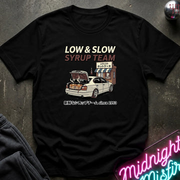 A black short-sleeve graphic T-shirt laid flat on a dark textured surface. The shirt features a printed design with the text 'LOW & SLOW SYRUP TEAM' above an illustration of a white car with its trunk open next to a small stand, plus Japanese text below.
