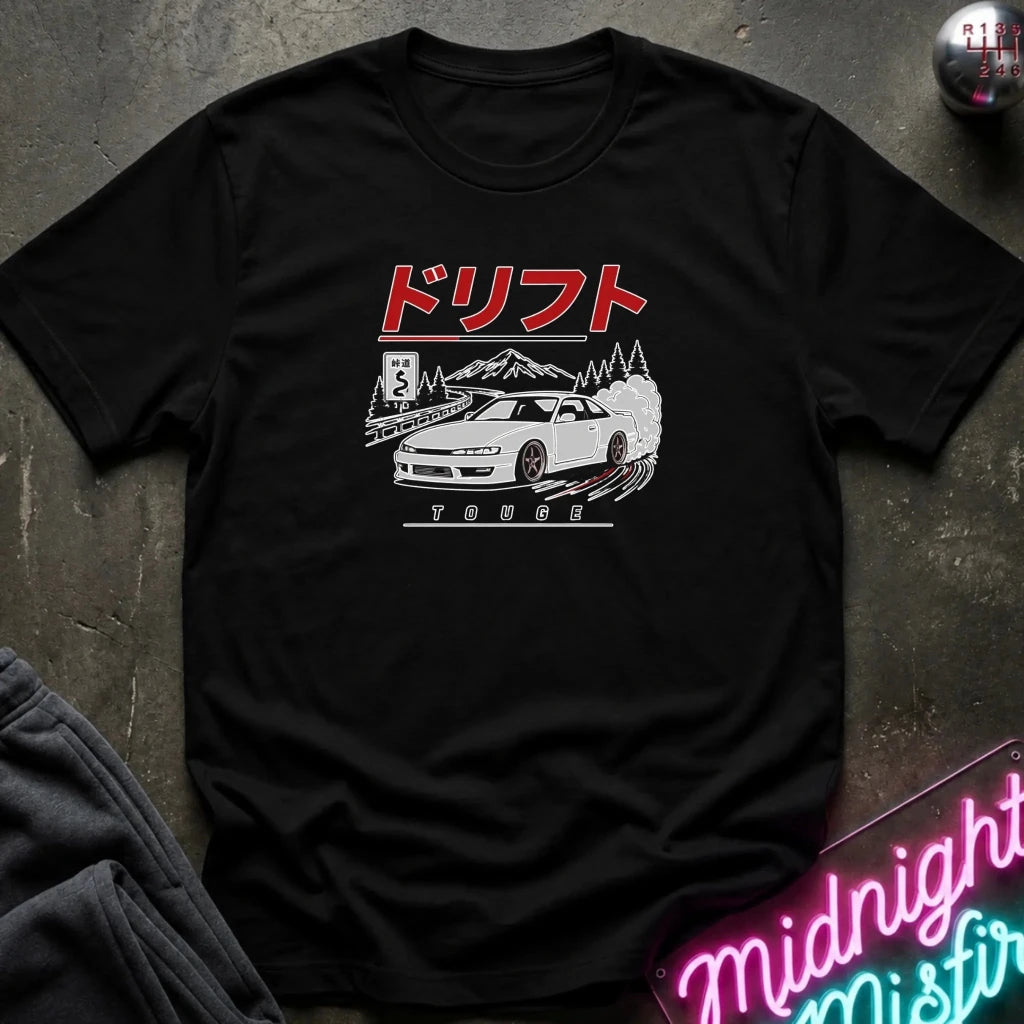 A black short-sleeve graphic T-shirt laid flat on a dark textured surface. The shirt features a white car illustration with red Japanese text above and the word 'TURBO' below.