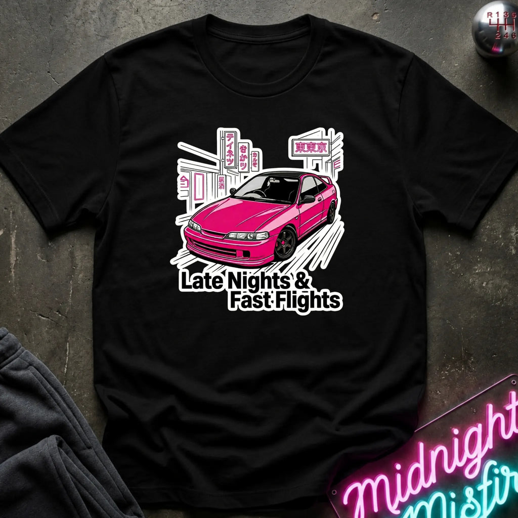 A black short-sleeve graphic t-shirt laid flat on a dark textured surface. The shirt features a graphic of a pink car in front of white buildings with the text 'Late Nights & Fast Flights'.