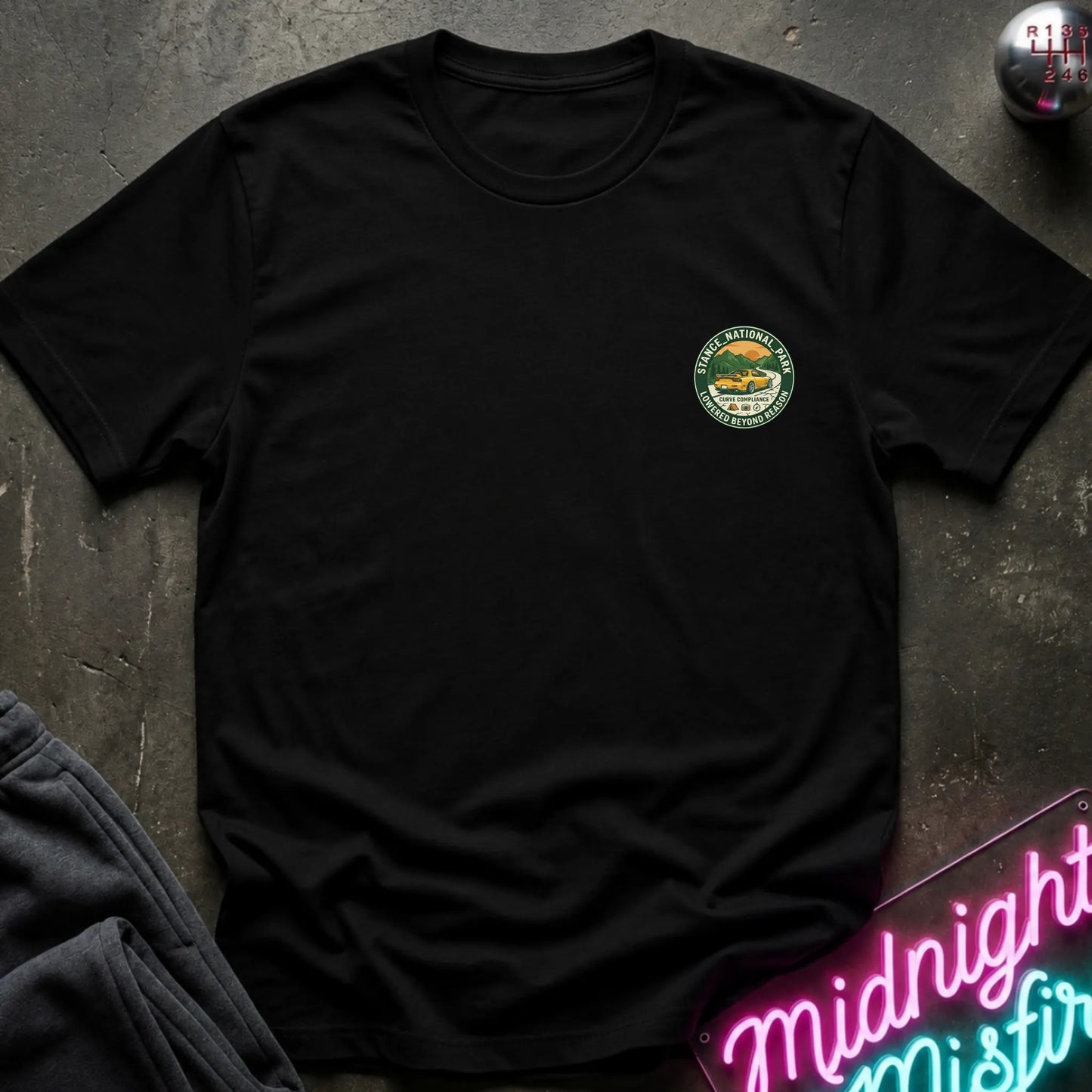 A black  crewneck t-shirt with a circular logo on the left chest, laid flat on a dark textured surface with a neon sign and dark pants in the background.