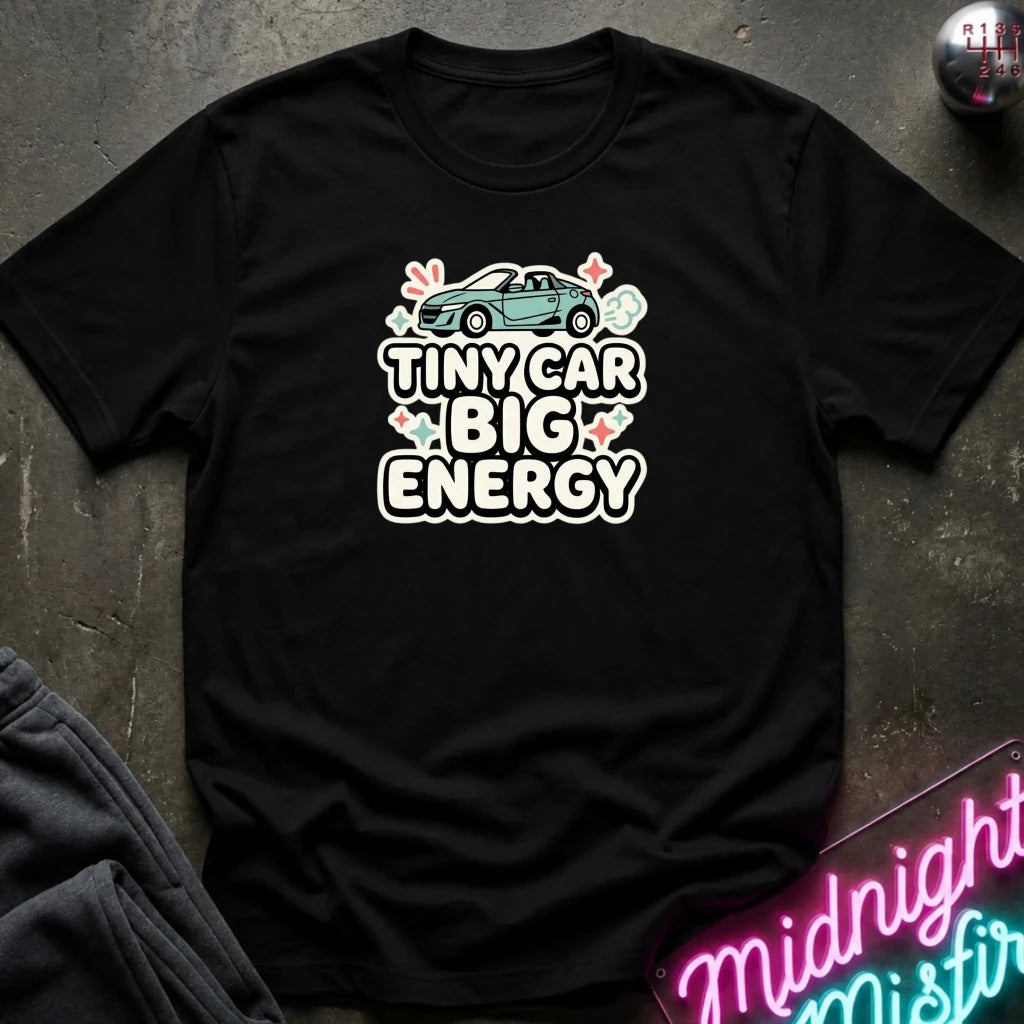 A black short-sleeve graphic T-shirt laid flat on a dark textured surface. The shirt features a graphic of a small teal car above the words 'TINY CAR BIG ENERGY' in white outlined letters with pink hearts and small floral accents.