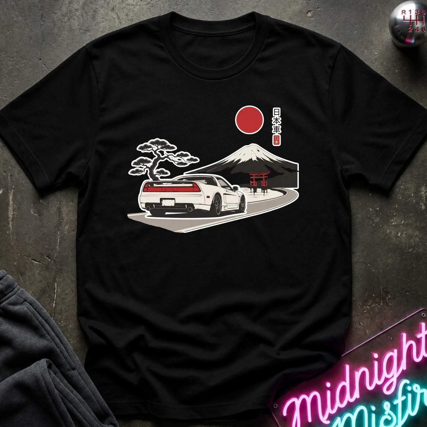 A black short-sleeve graphic t-shirt laid flat on a dark textured surface. The graphic features a white sports car on a road with a Mount Fuji background, a red sun, and Japanese text.