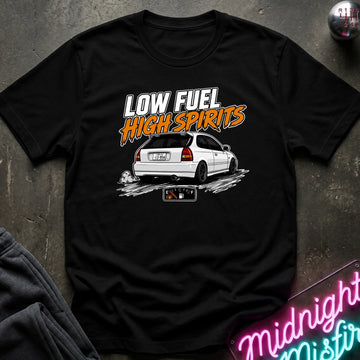 A black short-sleeve graphic t-shirt laid flat on a dark textured surface. The shirt features a white car with the text 'LOW FUEL HIGH SPIRITS' in white and orange.