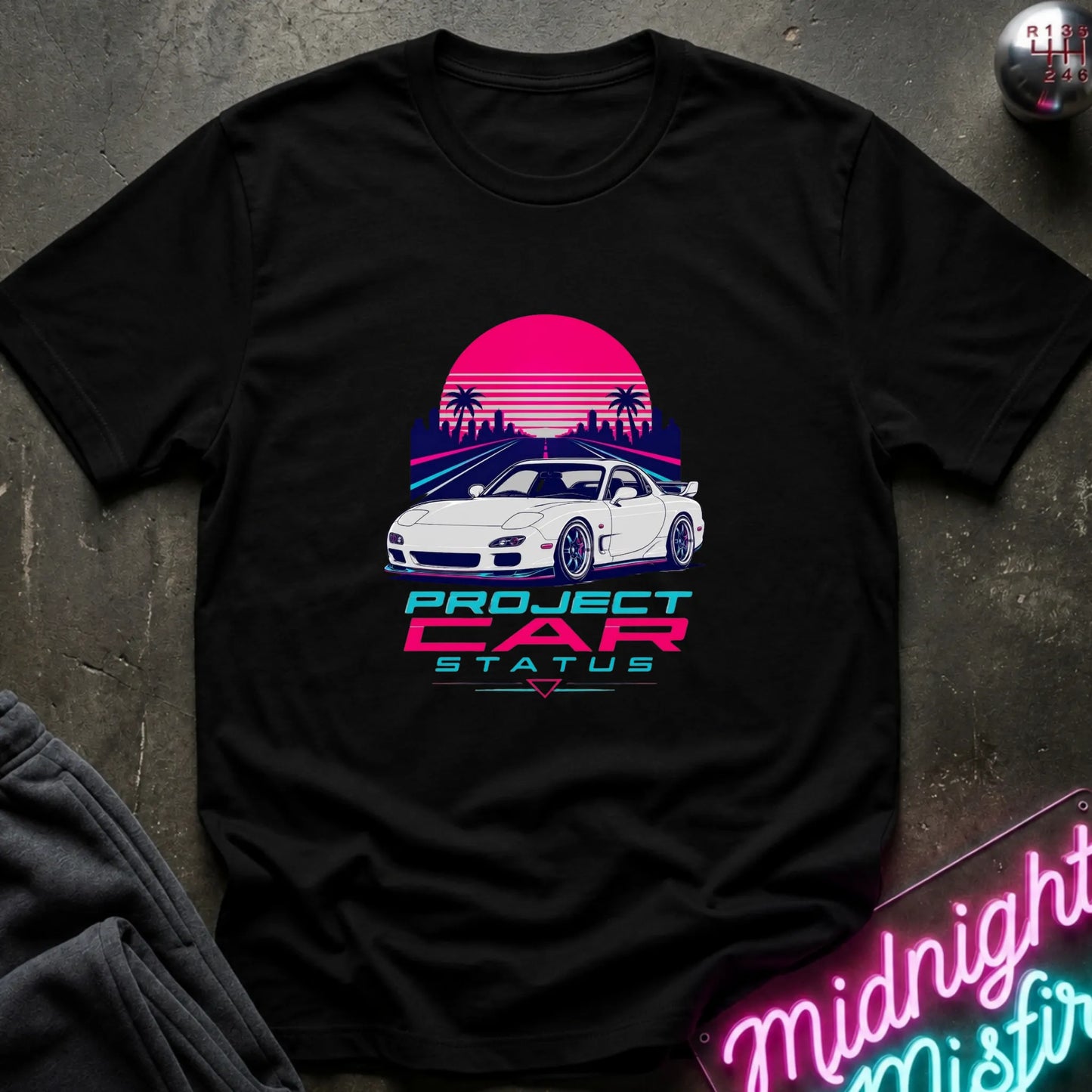 A black short-sleeve graphic T-shirt laid flat on a dark textured surface. The shirt features a retro-style print of a white sports car in front of a pink and purple sunset with palm trees, and the text 'PROJECT CAR STATUS' below.