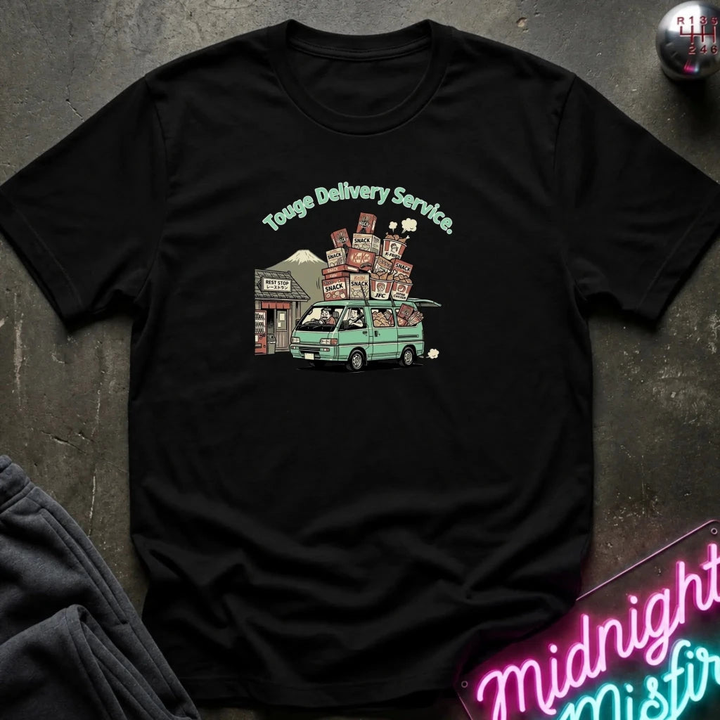 A black short-sleeve graphic t-shirt laid flat on a dark textured surface. The shirt features a colorful graphic of a green van carrying stacked packages, with the text 'Touge Delivery Service' above it.