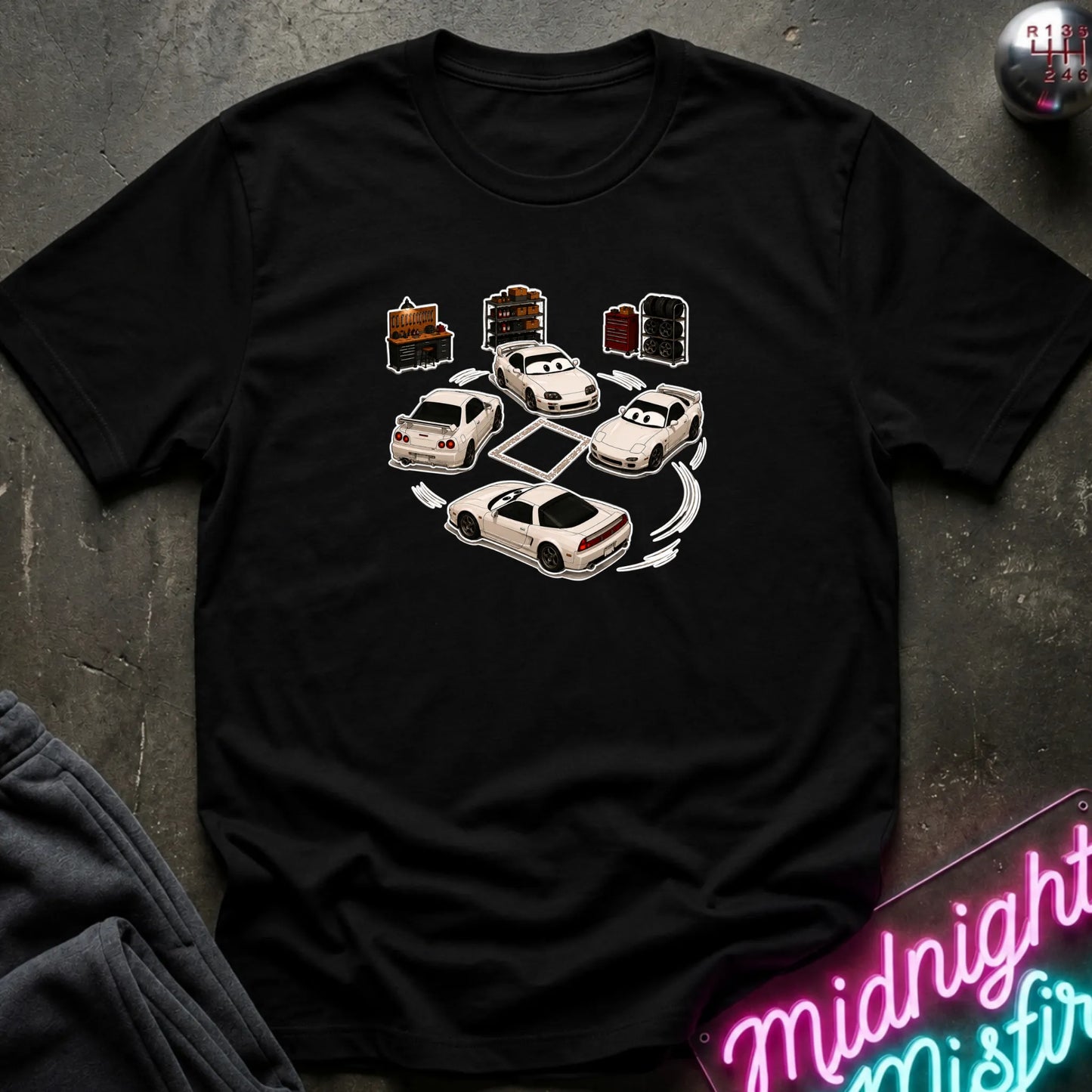 A black  short-sleeve t-shirt laid flat on a dark textured surface, featuring a graphic print of several white sports cars arranged in a circle with drift-like tire marks.